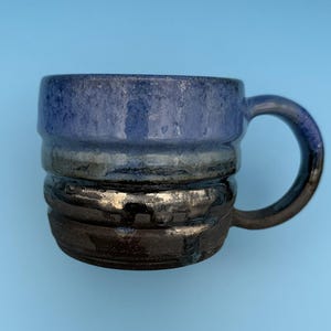 May include: A handmade ceramic mug with a dark blue, black, and silver glaze. The mug features a ridged design around the body and a rounded handle. The mug is set against a light blue background.