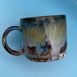 May include: A ceramic mug with a unique glaze in shades of blue, green, and brown. The mug has a rounded body and a sturdy handle. The glaze creates a marbled effect, adding visual interest to the handmade mug.