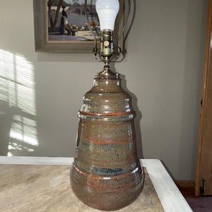 May include: A ceramic table lamp with a speckled brown and orange glaze. The lamp has a bulb and a metal frame. The lamp is on a wooden table. A framed painting hangs on the wall in the background.