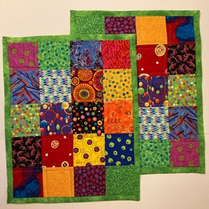 May include: Two colorful patchwork placemats with a green border. The placemats are made of various fabrics with different patterns, including polka dots, swirls, and geometric shapes. The placemats are a vibrant mix of red, yellow, green, blue, purple, and black.