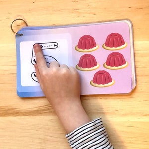 May include: A child's hand traces the number 5 on a blue and white card. The card has five red jelly cups on the right side.