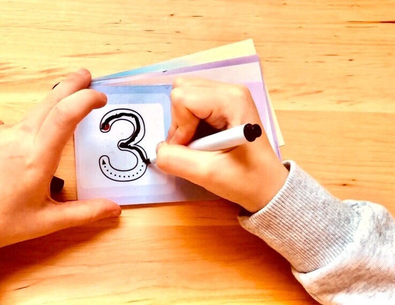 May include: A child's hand traces the number "3" on a white card with a dotted outline. The card is on top of a stack of colourful cards.