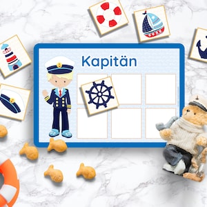 Puede incluir: Un tablero de juego azul y blanco con la palabra "Kapit&auml;n" impresa en &eacute;l. Hay seis casillas vac&iacute;as en el tablero. Hay varias tarjetas peque&ntilde;as con im&aacute;genes de un faro, un barco, un ancla, un capit&aacute;n, un volante y un gorro de marinero. Tambi&eacute;n hay un oso de peluche con un gorro de marinero y sosteniendo una pipa.