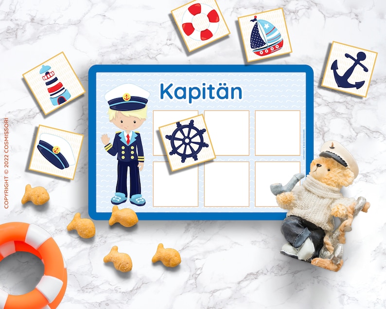 Puede incluir: Un tablero de juego azul y blanco con la palabra "Kapit&auml;n" impresa en &eacute;l. Hay seis casillas vac&iacute;as en el tablero. Hay varias tarjetas peque&ntilde;as con im&aacute;genes de un faro, un barco, un ancla, un capit&aacute;n, un volante y un gorro de marinero. Tambi&eacute;n hay un oso de peluche con un gorro de marinero y sosteniendo una pipa.