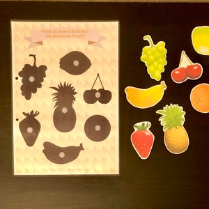 May include: A learning game for kids featuring a sheet with fruit silhouettes and matching fruit images. The text on the sheet reads "Finde zu jedem Schatten die passende Frucht!" which translates to "Find the matching fruit for each shadow!"