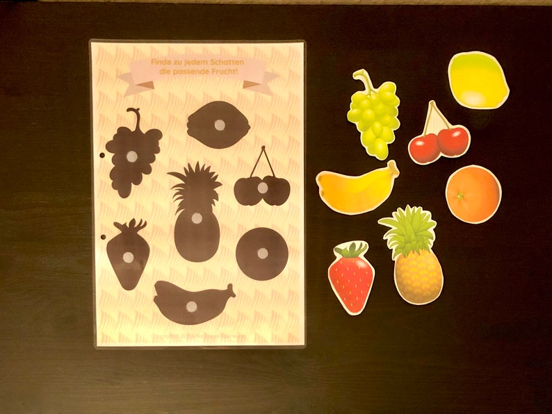 May include: A learning game for kids featuring a sheet with fruit silhouettes and matching fruit images. The text on the sheet reads "Finde zu jedem Schatten die passende Frucht!" which translates to "Find the matching fruit for each shadow!"