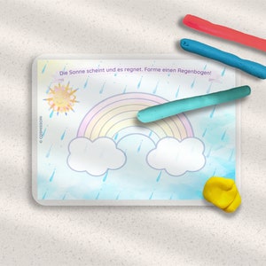 May include: A white card with a blue background and a rainbow, clouds, sun, and raindrops. The text on the card reads "Die Sonne scheint und es regnet.  Forme einen Regenbogen!"  There are three pieces of play dough in different colors: red, blue, and green.
