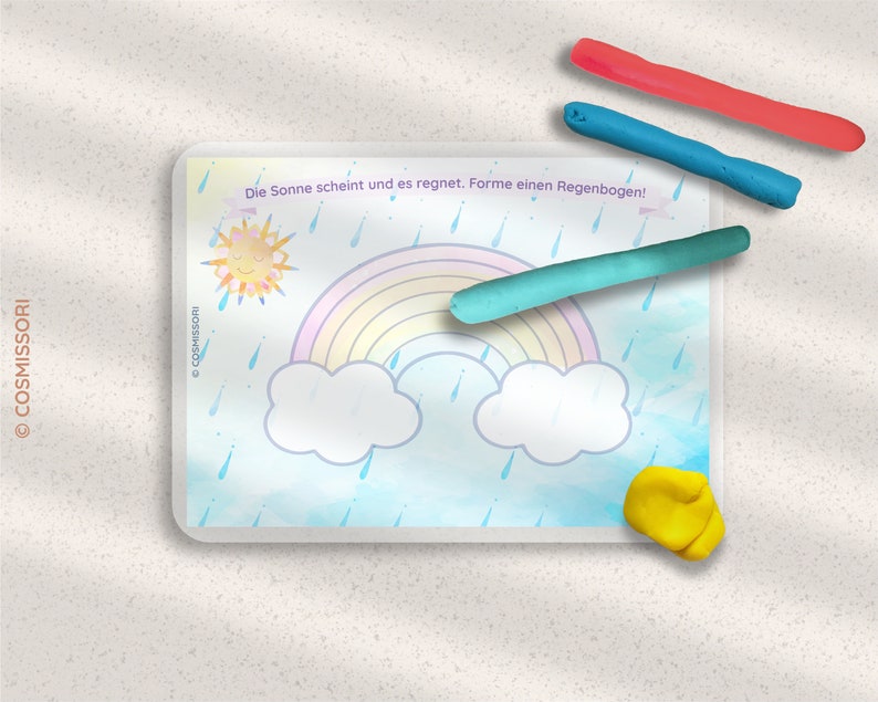 May include: A white card with a blue background and a rainbow, clouds, sun, and raindrops. The text on the card reads "Die Sonne scheint und es regnet.  Forme einen Regenbogen!"  There are three pieces of play dough in different colors: red, blue, and green.