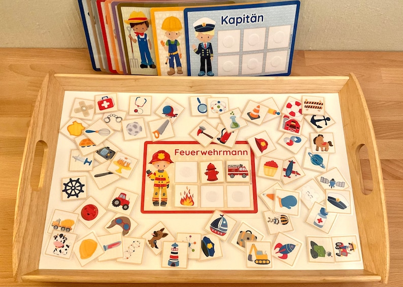 Puede incluir: Una bandeja de madera con una superficie blanca que contiene una variedad de tarjetas ilustradas de colores. Las tarjetas muestran im&aacute;genes de diferentes profesiones, incluyendo un bombero, un capit&aacute;n y un polic&iacute;a. Las tarjetas est&aacute;n dispuestas en un patr&oacute;n de cuadr&iacute;cula en la bandeja. La bandeja est&aacute; hecha de madera y tiene un acabado marr&oacute;n claro.