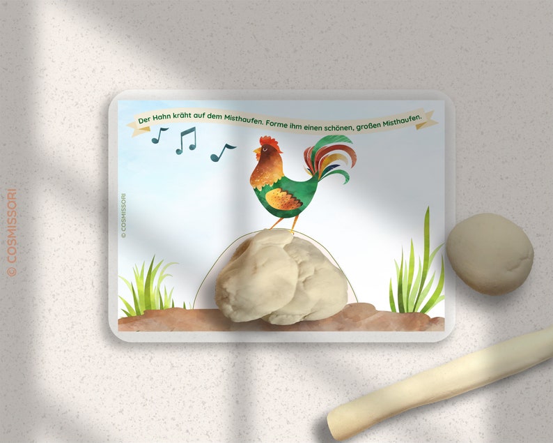 May include: A colorful illustration of a rooster crowing on a pile of dirt. The text on the card reads "Der Hahn kr&uuml;ht auf dem Misthaufen.  Forme ihm einen sch&ouml;nen, gro&szlig;en Misthaufen."
