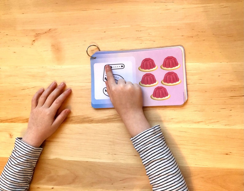 May include: A child's hand traces the number 5 on a blue and white card. The card has five red jelly cups on the right side.