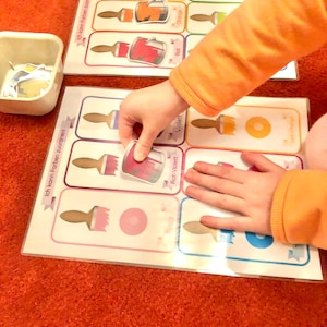 May include: A child's learning game with colorful paint cans and paintbrushes. The game includes the German words "Ich kann Farben zuordnen!" which means "I can match colors!"