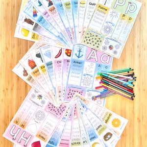 May include: A collection of colorful flashcards with German words and illustrations. The flashcards feature various objects, animals, and letters. The words on the flashcards include "Palme", "Anker", "Feuer", "Herz", "Kirsche", "Orange", and "Nest". A set of colorful markers is also included.