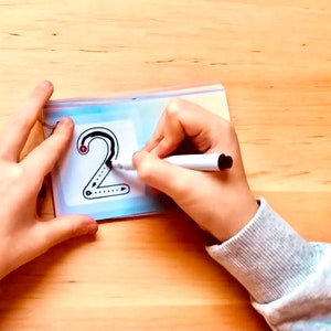 May include: A child's hand traces the number "2" on a clear plastic sheet with a white dry-erase marker. The number is outlined in black and has a red dot at the starting point.