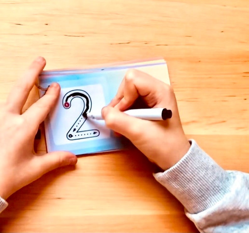 May include: A child's hand traces the number "2" on a clear plastic sheet with a white dry-erase marker. The number is outlined in black and has a red dot at the starting point.