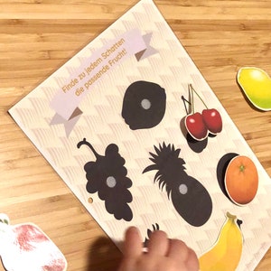 May include: A game for kids featuring a sheet of paper with black silhouettes of fruits and matching colorful fruit stickers. The text on the sheet reads "Finde zu jedem Schatten die passende Frucht!" which translates to "Find the matching fruit for each shadow!"