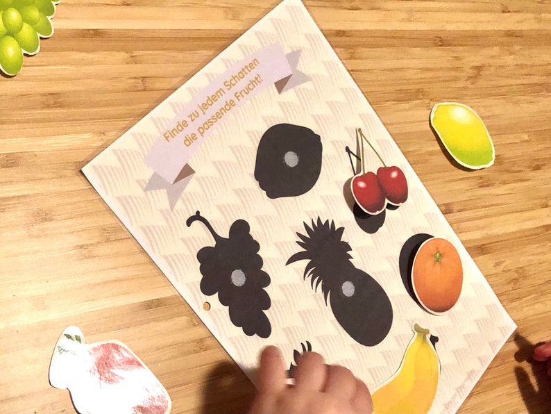 May include: A game for kids featuring a sheet of paper with black silhouettes of fruits and matching colorful fruit stickers. The text on the sheet reads "Finde zu jedem Schatten die passende Frucht!" which translates to "Find the matching fruit for each shadow!"