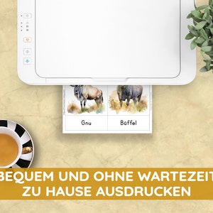 May include: A white printer printing animal flashcards with watercolor illustrations of a gnu and a buffalo. The flashcards have the words "Gnu" and "B&uuml;ffel" printed on them. A cup of tea in a black and white striped cup and saucer is in the lower left corner.