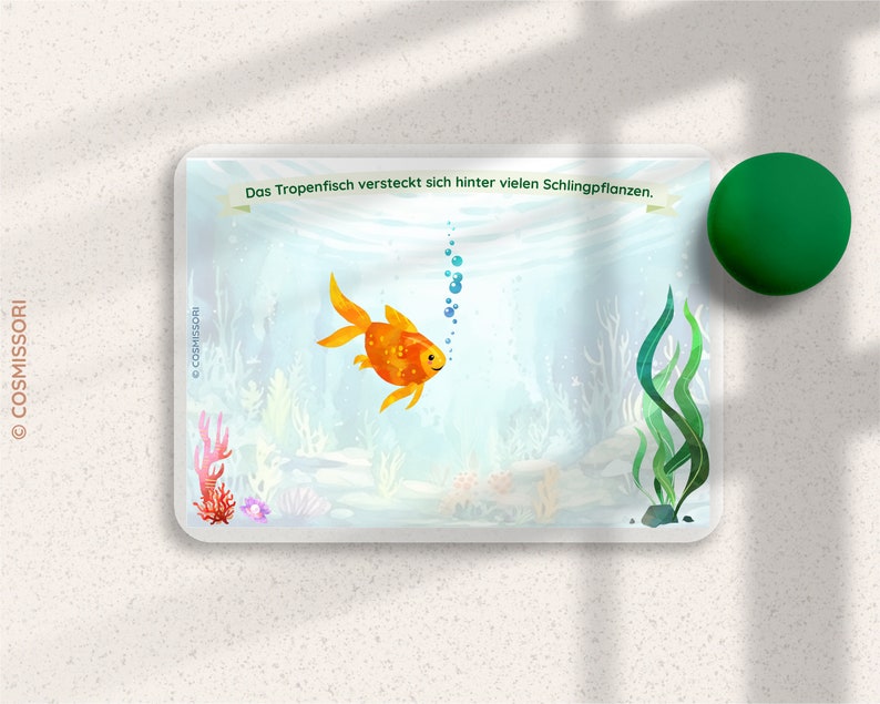 May include: A colorful illustration of a goldfish swimming in an underwater scene with green seaweed and bubbles. The text "Das Tropenfisch versteckt sich hinter vielen Schlingpflanzen." is at the top of the image.