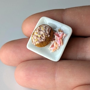 May include: A miniature square white plate holding a small brown textured food item topped with a light-coloured speckled mixture and a side portion of thin, shredded pink and white strips, presented as a tiny food replica for display purposes.