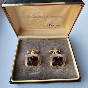 以下が含まれることがあります： 黒いベルベット張りの箱に入った、正方形の濃い茶色のガラスの石が付いた金色のカフスボタンのペア。箱は黒で金色の縁取りがされており、内蓋には「Admired the World Over」と「Anson Made in U.S.A.」の文字が印刷されています。