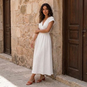 Può includere: Un abito bianco di lunghezza midi con maniche corte, scollo a V e vita aderente che scende in una gonna ampia. L'abito è abbinato a sandali marroni a punta aperta con tacco largo, offrendo un look semplice, pulito ed essenziale.