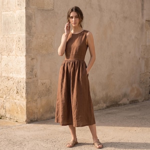 May include: A sleeveless midi dress in a solid brown color, featuring a round neckline, a fitted bodice, and a gathered skirt that falls below the knees. The dress has a simple, clean design with a defined waistline and side pockets.