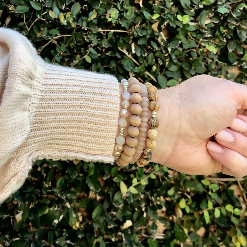May include: A set of five bracelets featuring wooden beads in shades of brown and beige, with some beads having a light pink hue. The bracelets are stacked on a person's wrist.