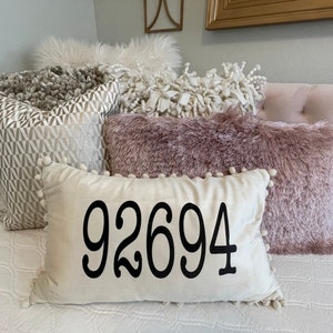 May include: A white throw pillow with black text that reads "92694" on a bed with a pink headboard and several decorative pillows.