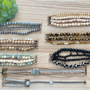May include: Eight different Apple Watch bands made with various beads and materials. The bands are in a variety of colors, including gray, brown, black, white, and gold. The bands are all layered and have a variety of textures.
