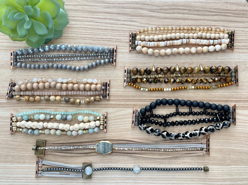 May include: Eight different Apple Watch bands made with various beads and materials. The bands are in a variety of colors, including gray, brown, black, white, and gold. The bands are all layered and have a variety of textures.