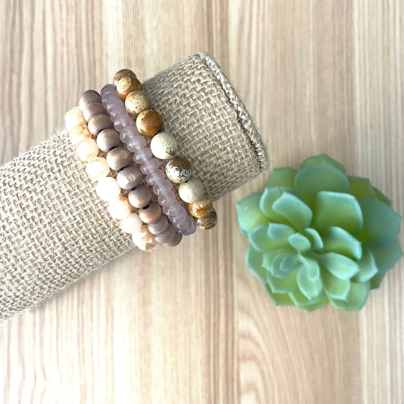 May include: A set of four bracelets made of natural stone beads in shades of brown, beige, and pink. The bracelets are stacked on a burlap fabric background.