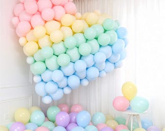 Pode incluir: Um grande arranjo de balões pastel em forma de coração em tons de rosa, amarelo, verde e azul. Os balões estão dispostos em um padrão degradê, com os balões rosa na parte superior e os balões azuis na parte inferior. Também há grupos menores de balões pastel no chão.