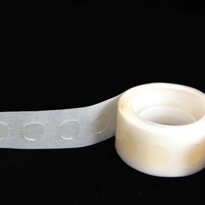 May include: A roll of clear adhesive dots on a white backing. The dots are designed for attaching lightweight items to surfaces.
