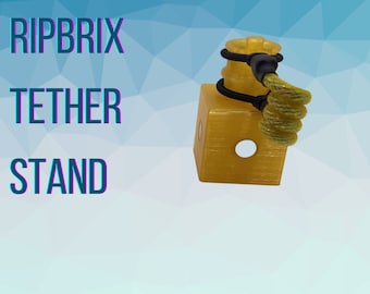 May include: A gold-colored plastic stand with a hole in the center and a black elastic band around the top. A yellow rope is attached to the band and is coiled around the stand. The text "RIPBRIX TETHER STAND" is visible in the background.