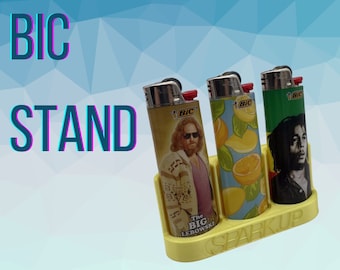 May include: A yellow BIC lighter stand holding three lighters with different designs. One lighter features a portrait of The Dude from The Big Lebowski, another has a lemon pattern, and the third has a portrait of Bob Marley.