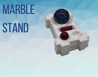 May include: White plastic marble stand with three compartments. The stand has a blue marble with white flecks in the top compartment, a red marble in the middle compartment, and an empty compartment on the bottom.