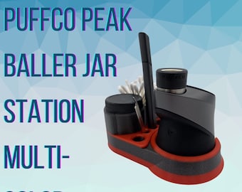 May include: A red and black Puffco Peak Baller Jar Station with a gray Puffco Peak vaporizer, a black tool, and a white cotton swab. The station has multiple compartments for storing accessories.