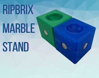 May include: Two 3D printed marble stands, one green and one blue, with a circular indentation on top. The stands are magnetic and have the text "RIPBRIX MARBLE STAND" printed on a background.