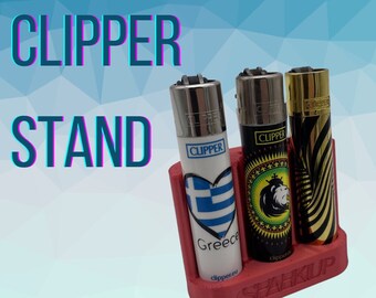May include: A red 3D printed stand for holding three lighters. The stand has the word "Sparkup" printed on it. The lighters are all different designs, one with a Greek flag, one with a lion, and one with a striped pattern. The lighters all have the word "Clipper" printed on them.