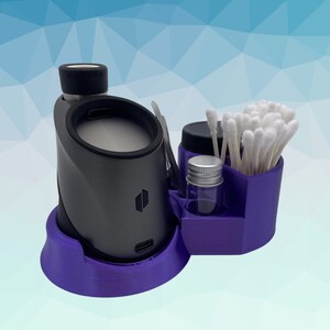 K&ouml;nnte beinhalten: Ein lilafarbener 3-teiliger Aufbewahrungsorganisator f&uuml;r einen Vaporizer. Der Organizer enth&auml;lt ein kleines Glasgef&auml;&szlig;, einen Wattest&auml;bchenhalter und eine Basis f&uuml;r einen Vaporizer. Der Vaporizer ist dunkelgrau und hat den Schriftzug "Puffco" aufgedruckt.