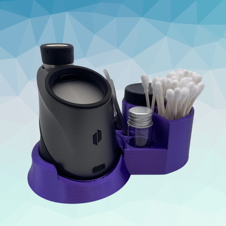 K&ouml;nnte beinhalten: Ein lilafarbener 3-teiliger Aufbewahrungsorganisator f&uuml;r einen Vaporizer. Der Organizer enth&auml;lt ein kleines Glasgef&auml;&szlig;, einen Wattest&auml;bchenhalter und eine Basis f&uuml;r einen Vaporizer. Der Vaporizer ist dunkelgrau und hat den Schriftzug "Puffco" aufgedruckt.