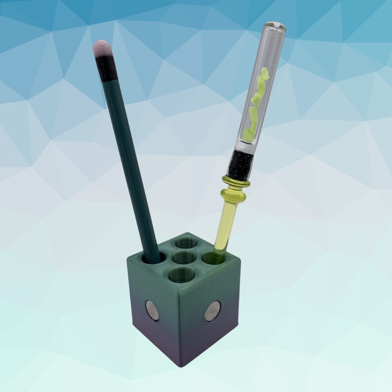 Puede incluir: Un porta l&aacute;pices y bol&iacute;grafos impreso en 3D de color verde y morado con seis ranuras. Un tubo de vidrio verde con un l&iacute;quido amarillo est&aacute; en una ranura y un l&aacute;piz negro en otra.