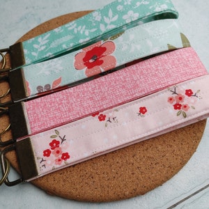 May include: Four fabric keychains with floral patterns and metal key rings. The keychains are in shades of pink and green with white floral designs.