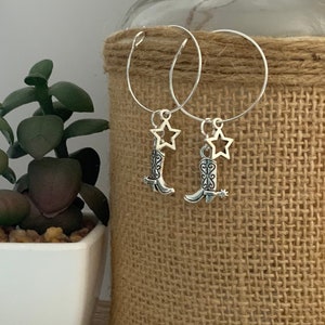 May include: Silver hoop earrings with cowboy boot and star charms. The earrings feature small, detailed boot pendants with spurs and a star-shaped charm. The earrings are displayed against a neutral background.