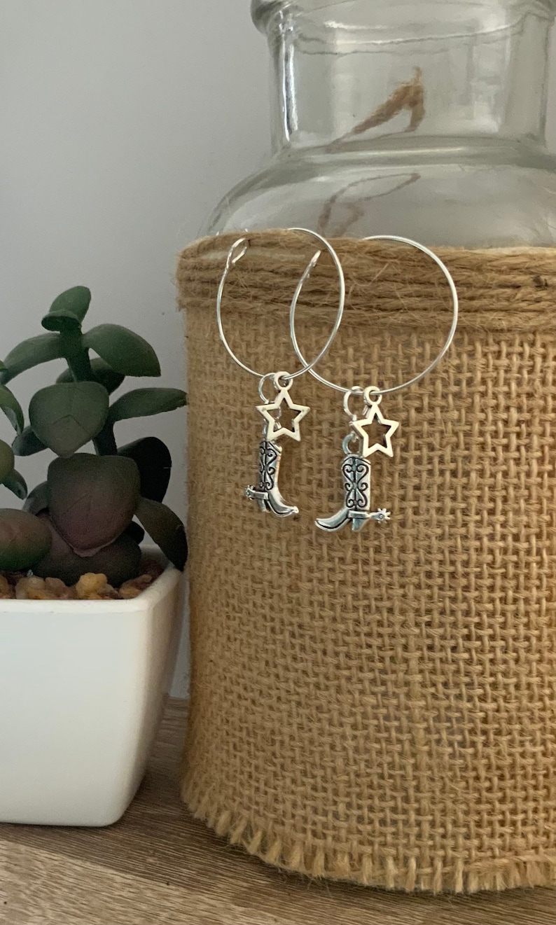 May include: Silver hoop earrings with cowboy boot and star charms. The earrings feature small, detailed boot pendants with spurs and a star-shaped charm. The earrings are displayed against a neutral background.