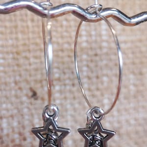 May include: Silver-toned hoop earrings with a Western theme. Each earring features a star charm above a detailed cowboy boot pendant. The earrings are displayed against a neutral, textured background.