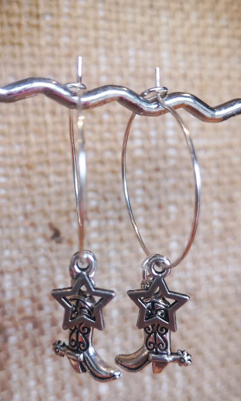 May include: Silver-toned hoop earrings with a Western theme. Each earring features a star charm above a detailed cowboy boot pendant. The earrings are displayed against a neutral, textured background.