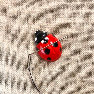 May include: A red and black ladybug with white spots is being threaded with a needle on a beige fabric background.