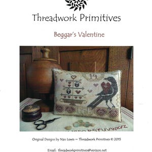 Puede incluir: Un cojín rectangular bordado con un pájaro negro que sostiene un corazón rojo, un soporte de niveles con corazones y el número 14. El texto Threadwork Primitives, Beggar's Valentine y Original Designs by Nan Lewis ~ Threadwork Primitives © 2015 es visible.
